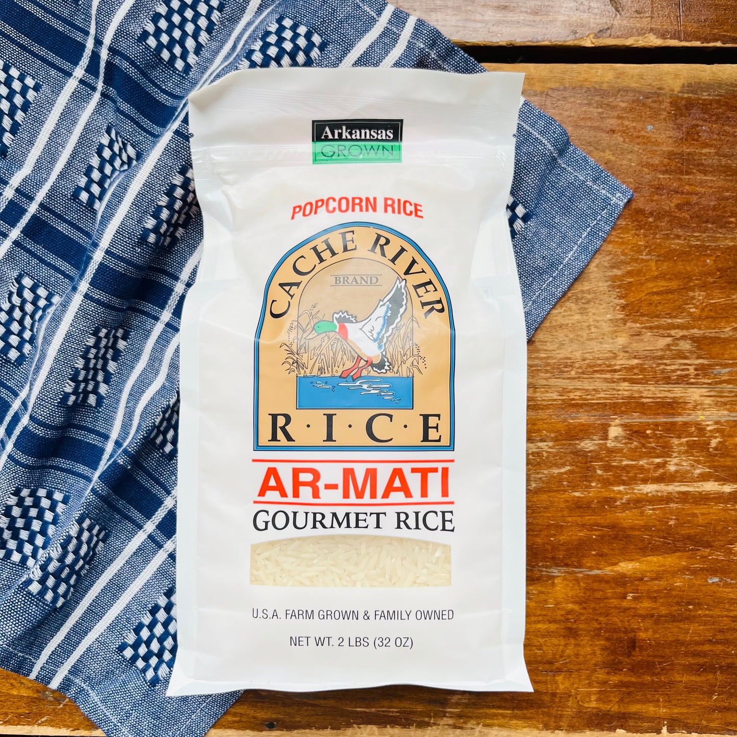 A bag of Cache River White Basmati Rice, labeled as 'AR-MATI Gourmet Rice', placed on a wooden surface with a blue patterned cloth.