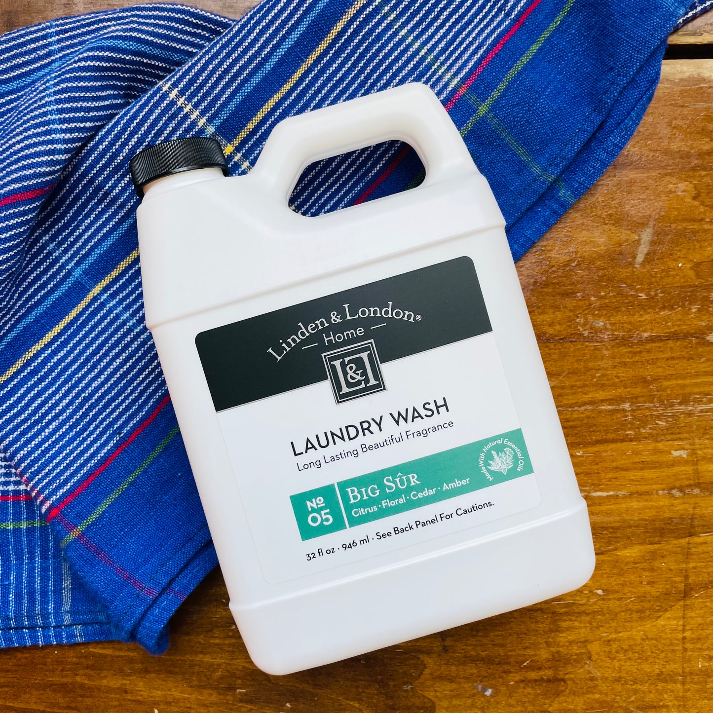 A bottle of Linden & London laundry detergent with a label and a green cap, resting on a wooden surface with a blue patterned fabric beside it. Laundry wash that smells of citrus, floral, cedar and amber