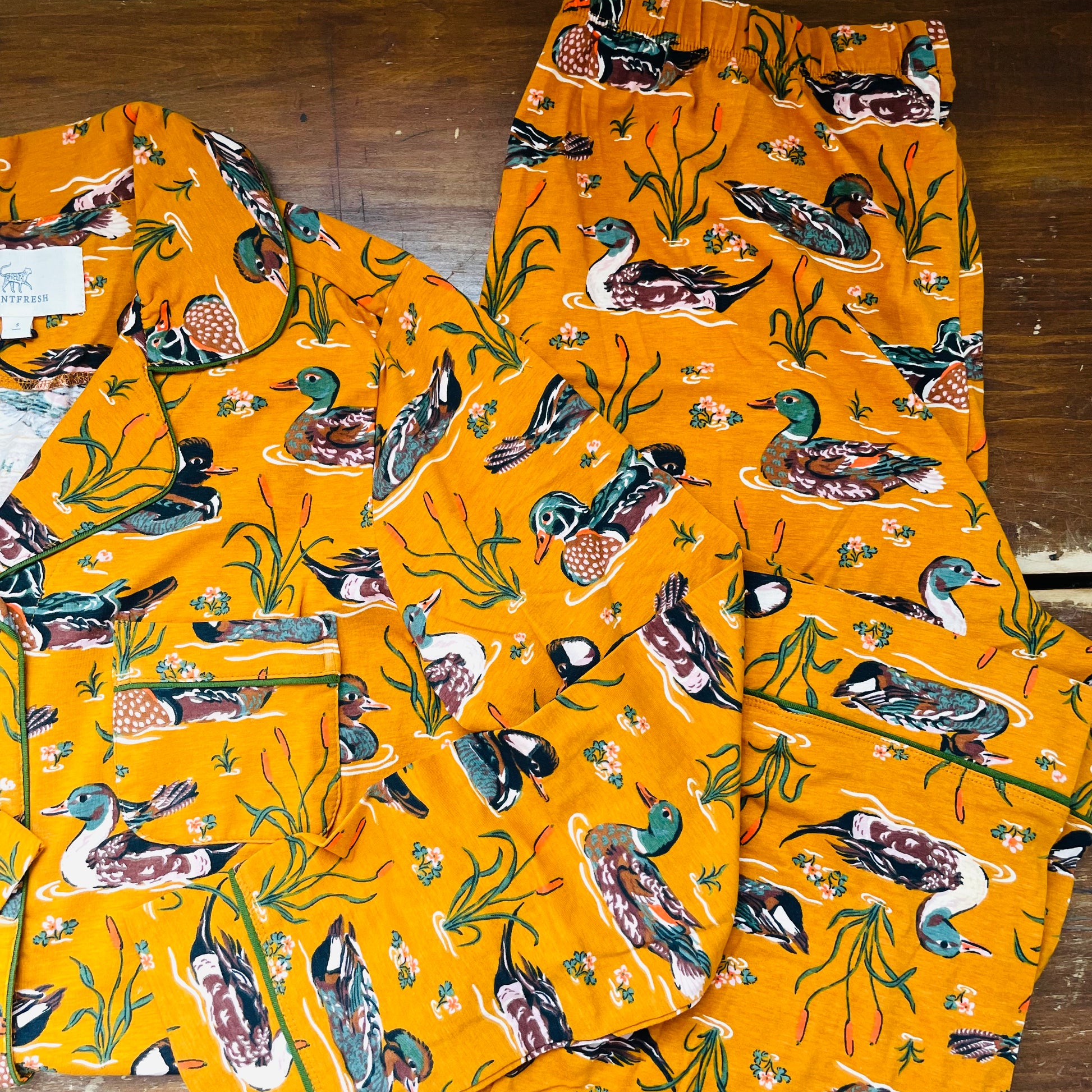 A two-piece pajama set featuring a bright orange-yellow background printed with a detailed pattern of ducks, geese, and water plants. The set includes a long-sleeve top with a collar and matching pants with an elastic waistband, laid flat on a dark wooden surface.