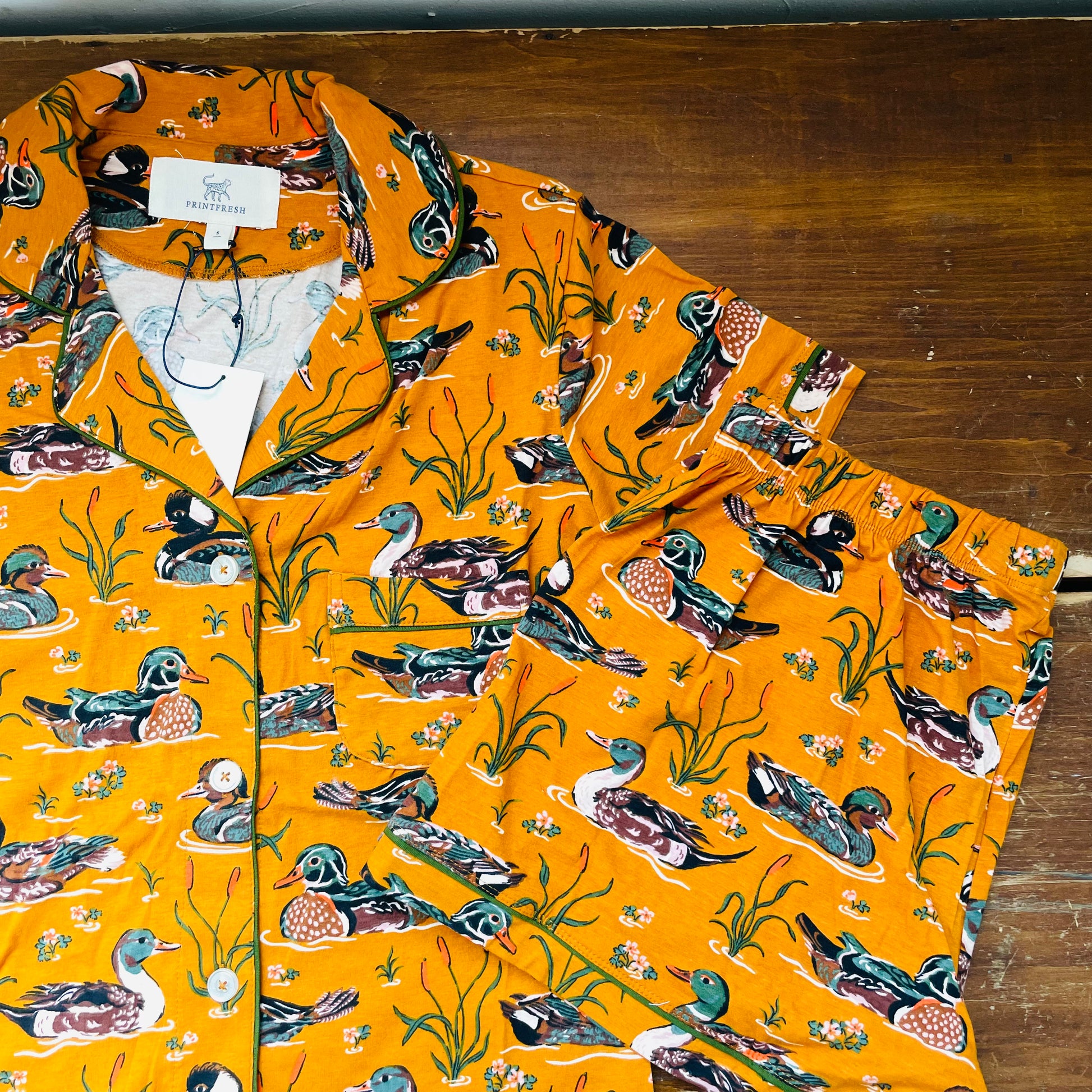 A bright orange button-up shirt with an all-over print of ducks, fish, and reeds, laid flat on a dark wooden surface. The brand name 'The Duckery' is visible on the inner neck label.