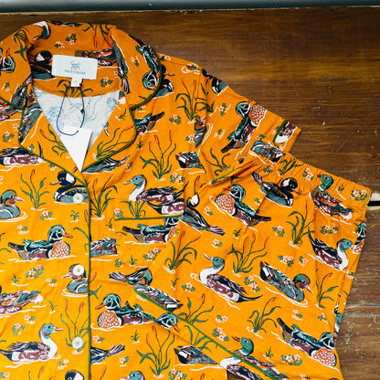 A bright orange button-up shirt with an all-over print of ducks, fish, and reeds, laid flat on a dark wooden surface. The brand name 'The Duckery' is visible on the inner neck label.
