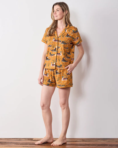 A woman modeling a matching two-piece pajama set with a mustard yellow background and a black, white, and red abstract pattern, standing against a plain white wall.