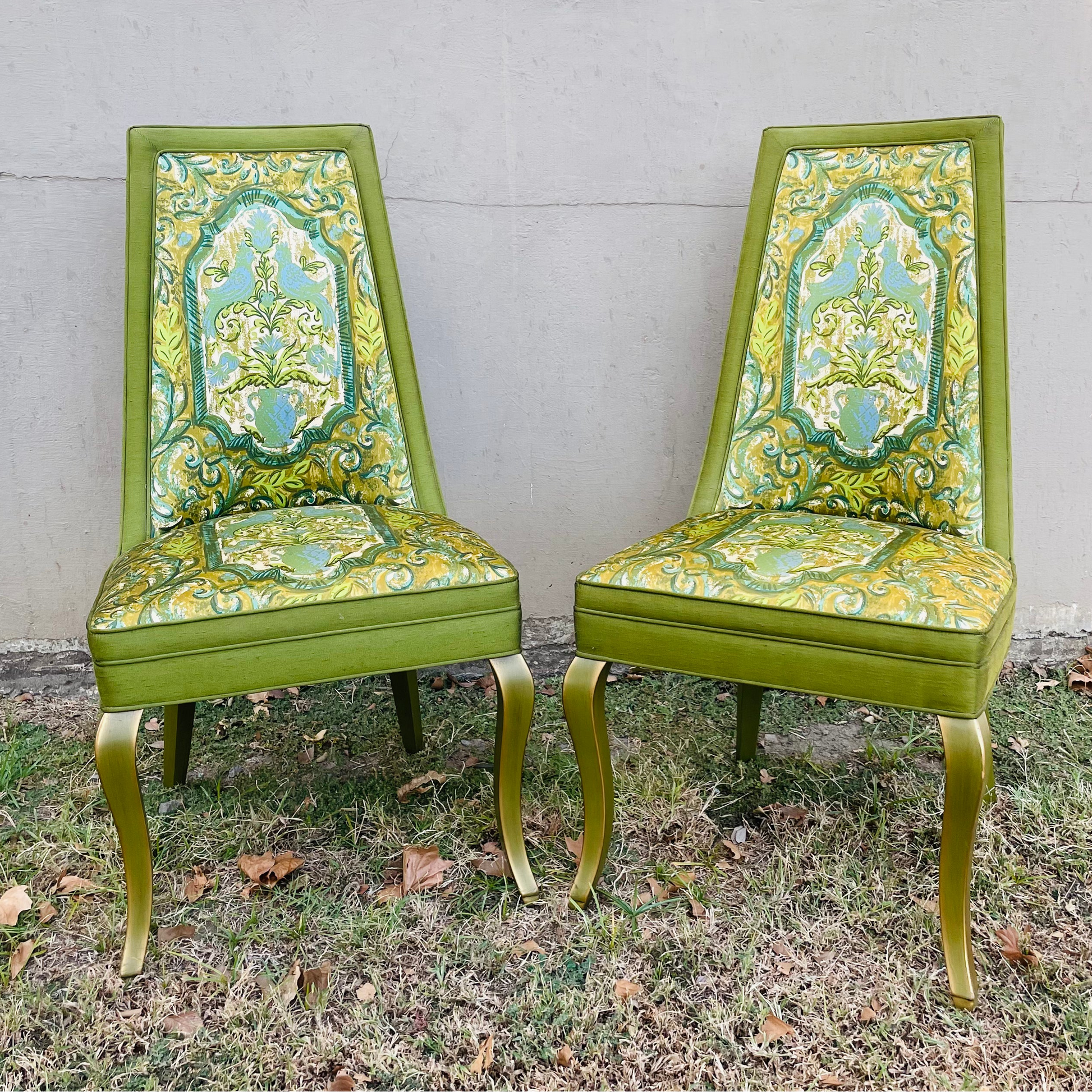 A pair of high-back dining chairs with olive green frames and ornate floral patterned upholstery in green, yellow, and blue, set on grass against a concrete wall.