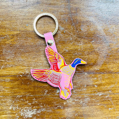 A multicolored keychain featuring a embossed flying duck design with a gold plated ring, on a wooden surface.