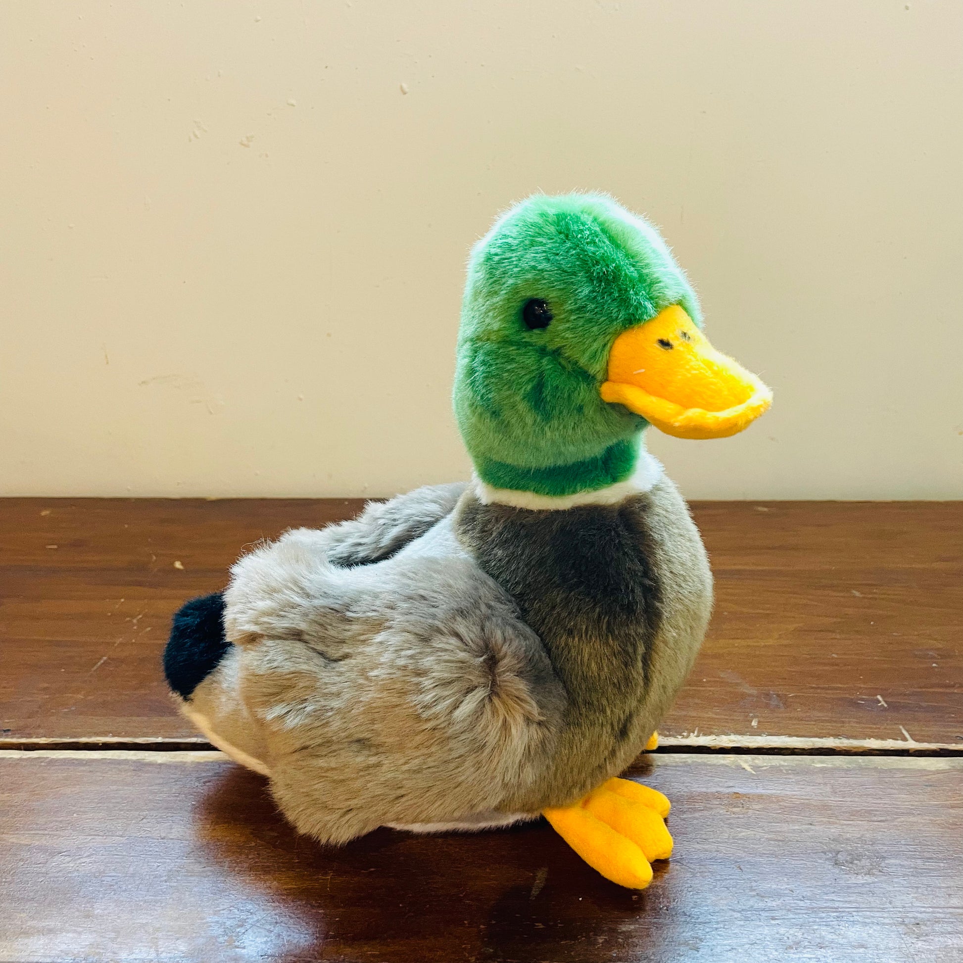 A plush Mallard Drake toy with a green head, gray body, and yellow beak and feet, sitting on a wooden surface.