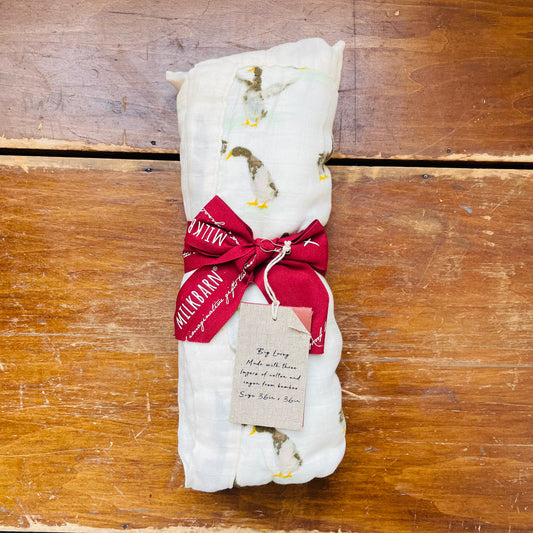 A white three-layer muslin blanket with a duck print, tied with a red ribbon and adorned with a name tag, on a wooden surface.