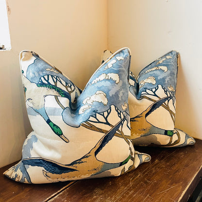 Two blue square throw pillows with a pattern of flying ducks against a tree silhouette background, placed on a surface.
