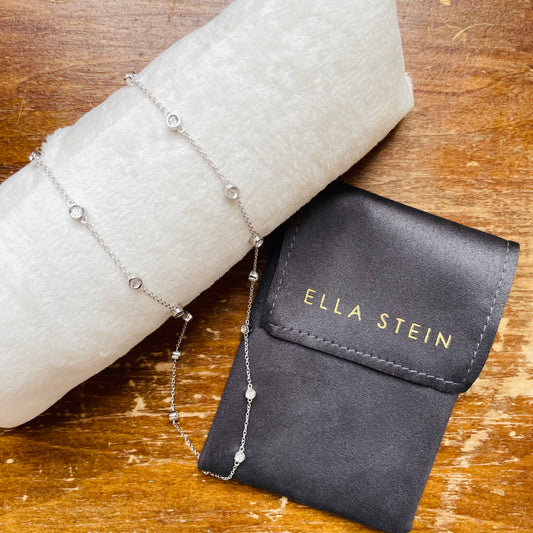 A delicate silver chain bracelet with small clear gemstone accents, displayed on a white textured tube next to an Ella Stein black velvet pouch on a wooden surface.
