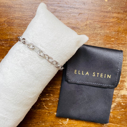 A silver chain bracelet displayed on a white fabric jewelry pillow next to an Ella Stein brand pouch on a wooden surface.