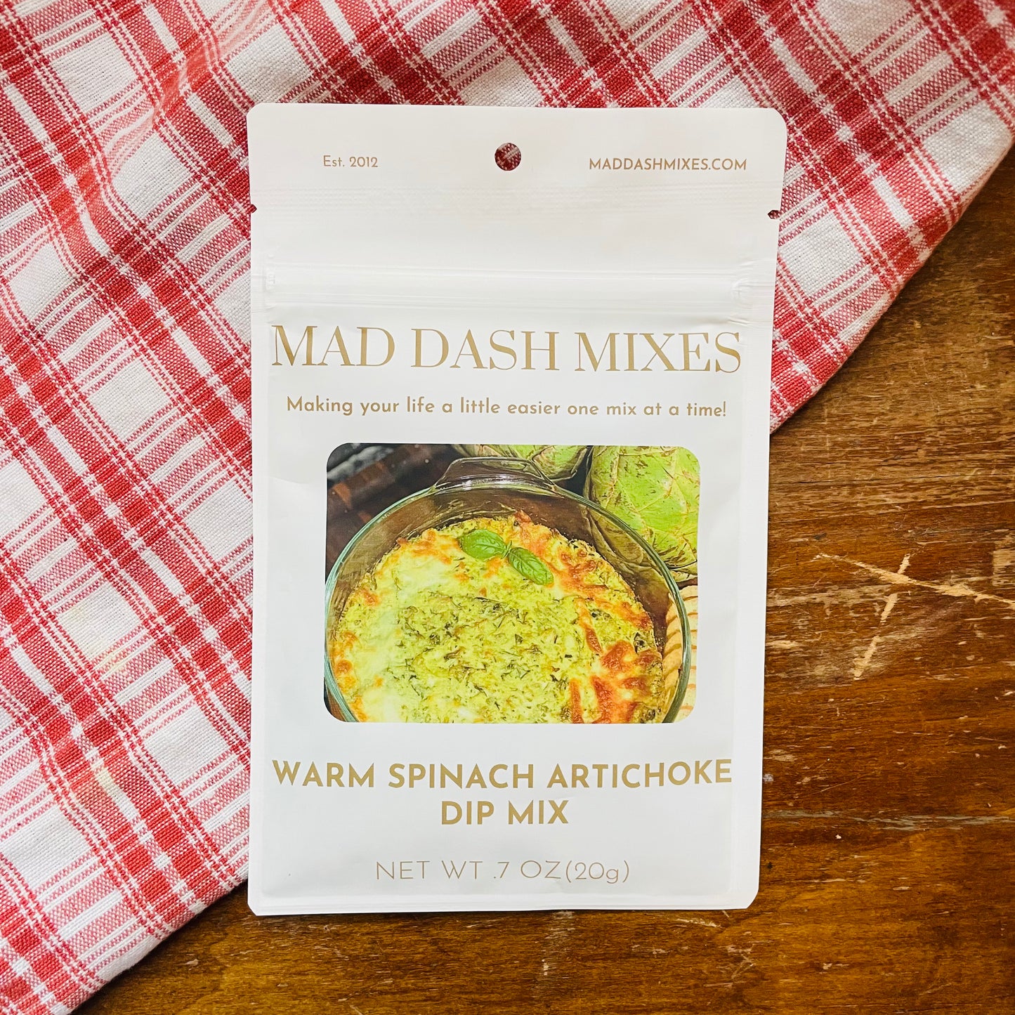 A package of Mad Dash Mixes Warm Spinach Artichoke Dip Mix, placed on a wooden surface with a red and white checkered cloth.