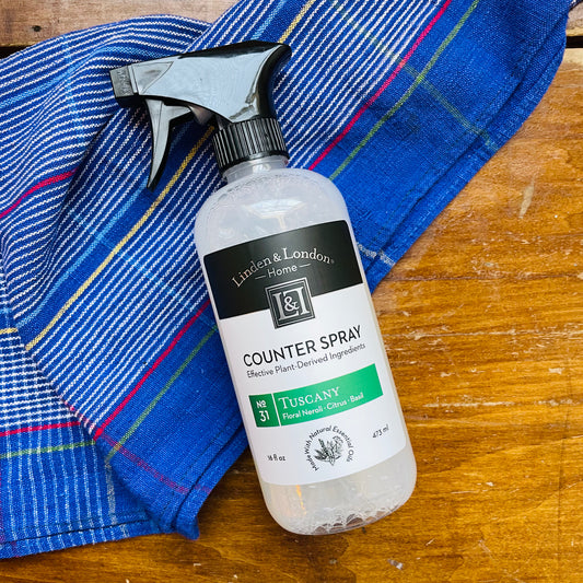 A white bottle of Counter Spray by Linden & London placed on a wooden surface with a blue striped cloth beside it. Counter spray that smells like floral, neroli, citrus and basil. 