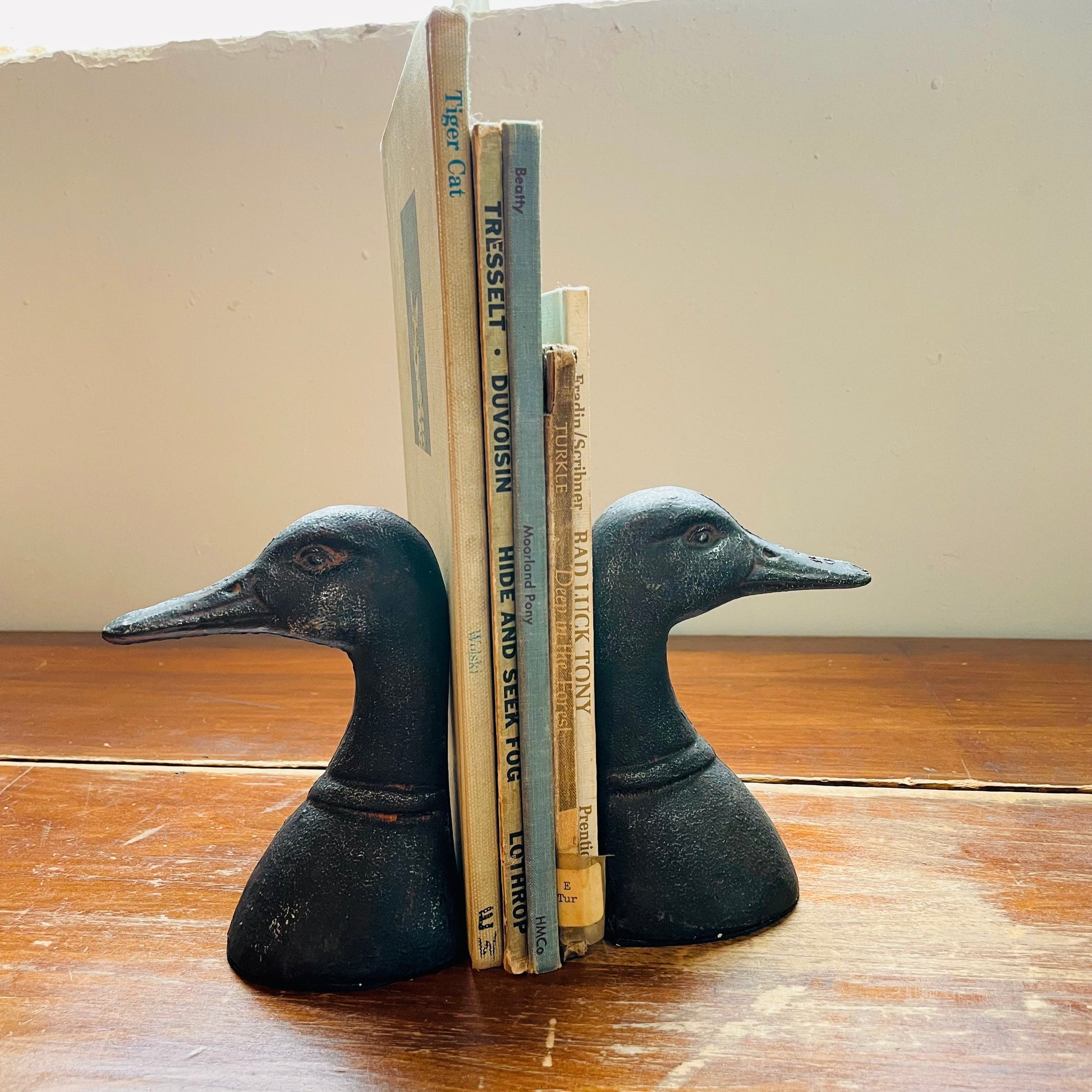 Two black cast iron duck head bookends placed on a wooden surface, holding up a couple of books.