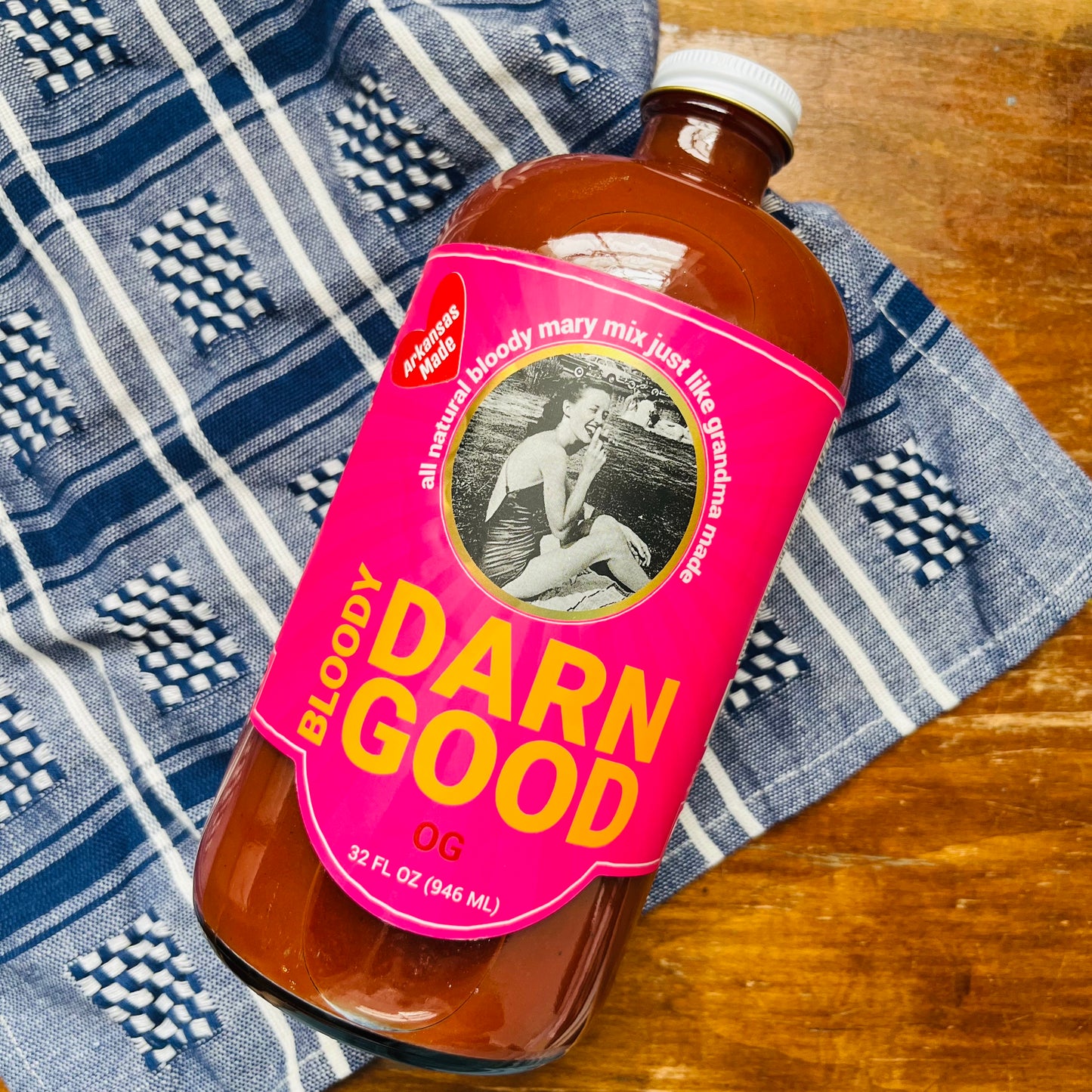 A bottle of OG Bloody Mary Mix on a wooden surface, with a blue and white patterned cloth in the background.