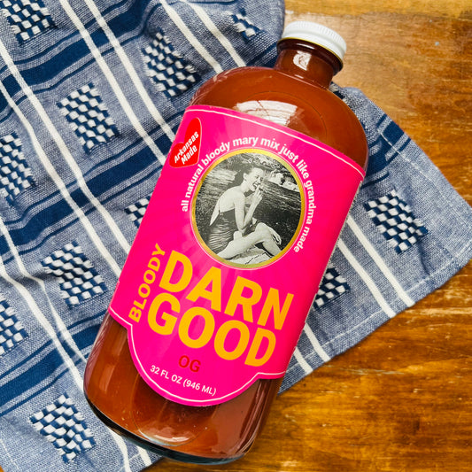 A bottle of OG Bloody Mary Mix on a wooden surface, with a blue and white patterned cloth in the background.